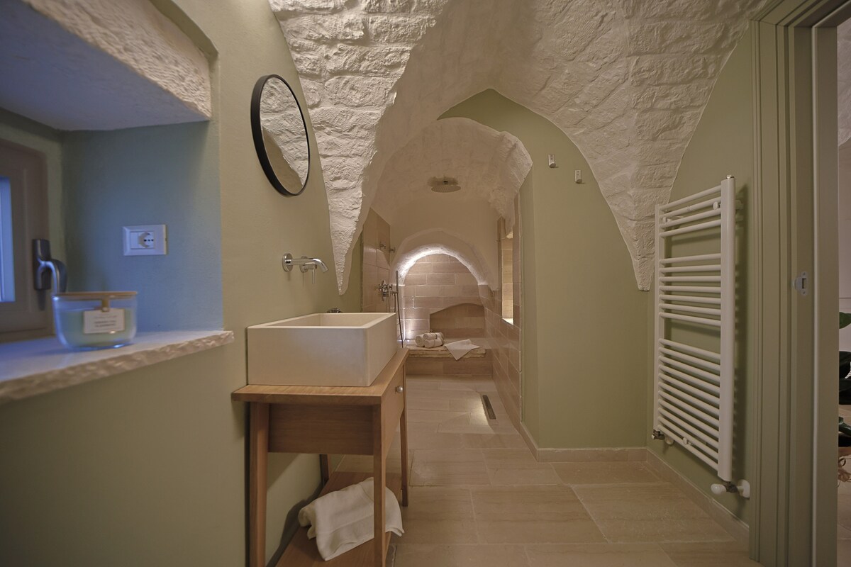 A spacious bathroom features stone arched ceilings and soft green walls, enhancing the natural light. A wooden vanity with a rectangular sink stands beside a large framed mirror. A towel is neatly placed on the floor, and a heated towel rack is mounted on the wall.