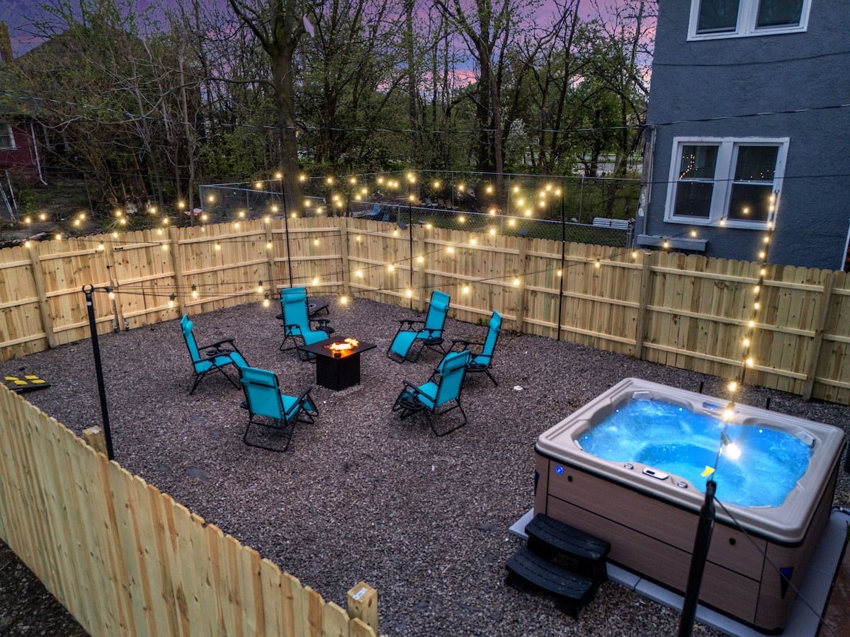 A private fenced backyard is illuminated by string lights, creating a relaxed atmosphere. A hot tub sits in one corner, while a fire pit is surrounded by several lounge chairs. The gravel surface provides a comfortable setting for outdoor gatherings.