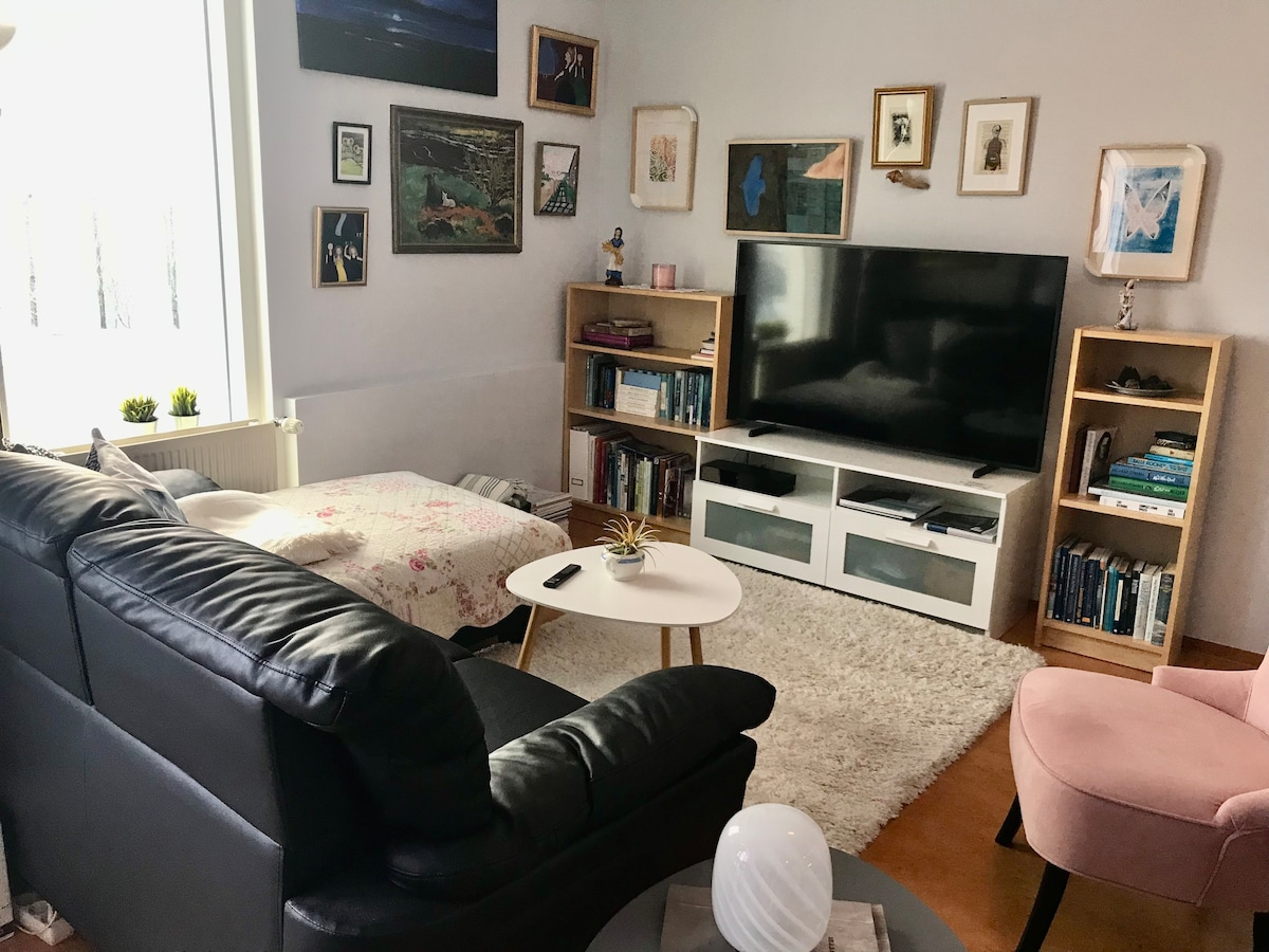 An open living room is depicted, featuring a sleek black couch and a decorative coffee table. A large television is positioned on a modern white cabinet. The walls are adorned with framed artwork, while bookshelves display various books. A plush rug anchors the seating area.