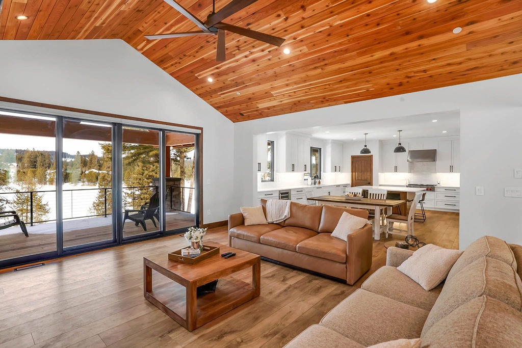 A spacious living area features a light-colored sofa and a wooden coffee table, complemented by a rustic ceiling with exposed beams. Large glass doors open to a balcony, enhancing natural light. The adjacent dining space includes a wooden table surrounded by white chairs.