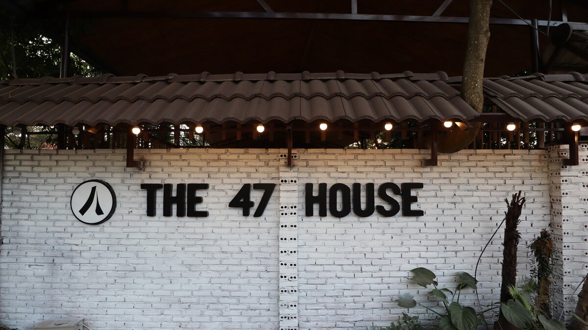 The exterior wall of The 47 House is featured, showcasing a white brick design. The name 'THE 47 HOUSE' is displayed in bold, black letters. Soft lighting from overhead bulbs contributes to a welcoming atmosphere, with greenery visible in the foreground.