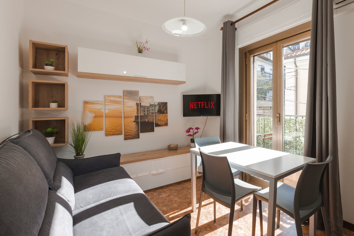 A modern living area features a comfortable dark grey sofa and a dining table with four chairs. A flat-screen television displaying the Netflix logo is mounted on the wall. Natural light enters through the glass door leading to a balcony, accentuated by decorative wall art.