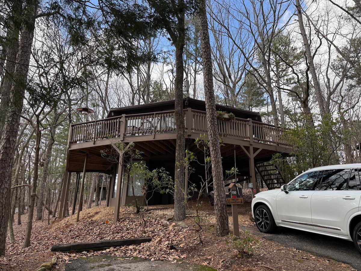 A charming cabin sits elevated on stilts, surrounded by leafless trees, showcasing a spacious deck that wraps around the structure. The exterior features natural wood tones, and a parked vehicle is visible nearby on a gravel path.