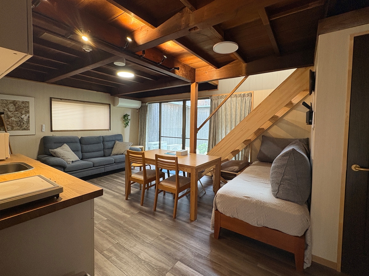 An open living space features a comfortable sofa and a wooden dining table surrounded by chairs. Natural light streams through the window adorned with light curtains, while a staircase leads to the upper floor. A versatile futon is positioned beside the dining area.