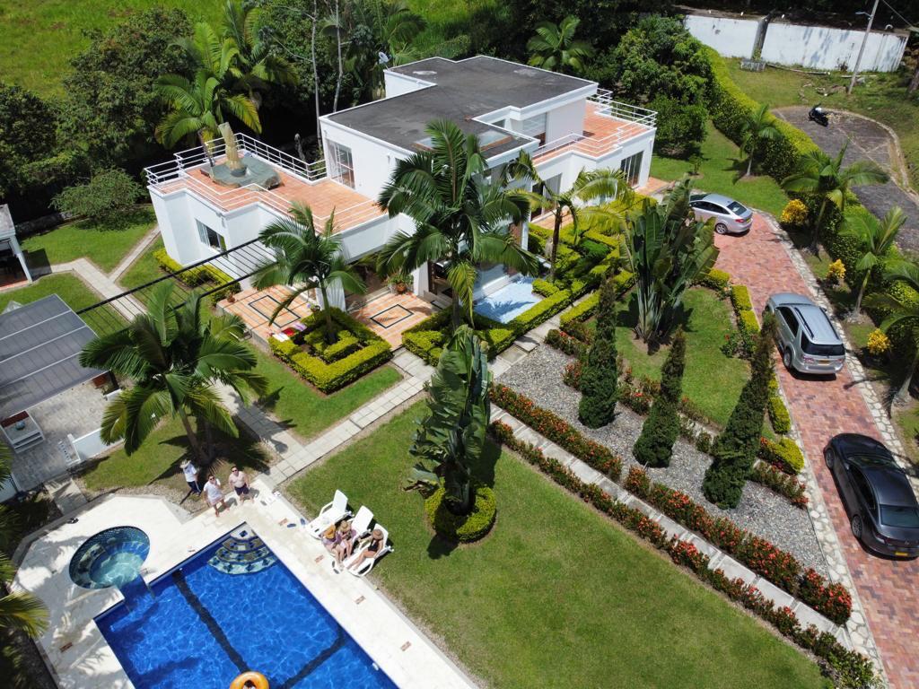 An aerial view showcases a spacious house surrounded by lush gardens and palm trees. A large pool features a jacuzzi, while a vibrant garden landscape adds color. Several cars are parked in the driveway, highlighting the accessible layout of the property.