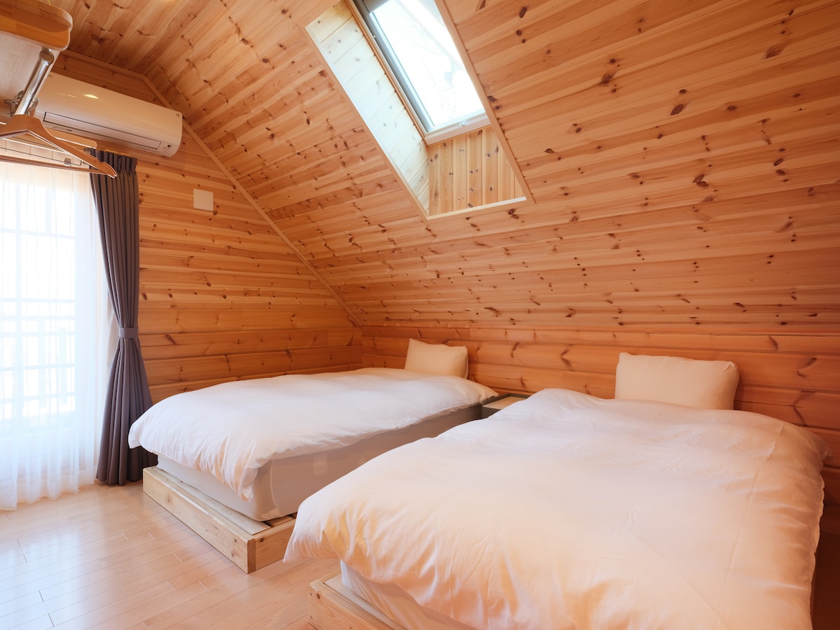 シンプルな2台のシングルベッドが配置され、透け感のあるカーテンやグレーの遮光カーテン、木に馴染むエアコンなど、全体的にナチュラルで穏やかな配色と素材で統一され、静かで心地よい雰囲気のベットルームです。