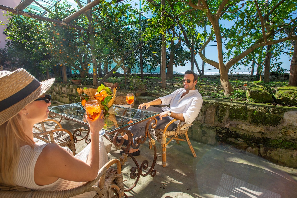 A serene outdoor seating area features a glass table surrounded by wicker chairs. Lush greenery frames the space, providing a natural backdrop. Two beverages are placed on the table, reflecting the leisurely atmosphere of the setting.