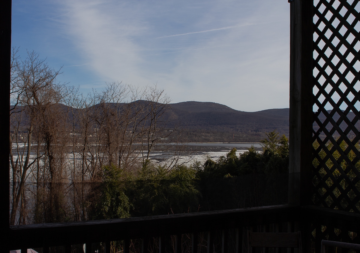 A serene view of the Hudson River is framed by a wooden lattice railing. Majestic mountains rise in the background, their silhouettes soft against a clear sky, while the river's surface reflects the natural beauty of the surrounding landscape.