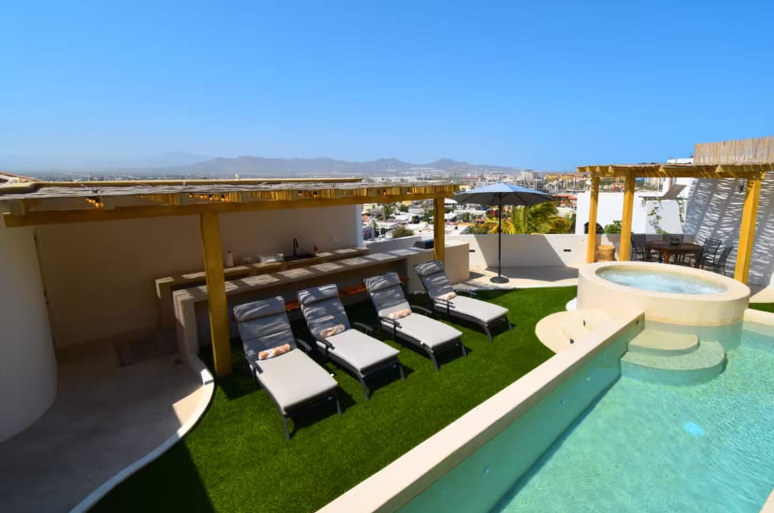 A sun-soaked terrace showcases a refreshing pool and a bubbling jacuzzi. Six lounge chairs are arranged on lush green turf, accompanied by a shaded umbrella. The view captures the surrounding landscape and distant mountains, enhancing the outdoor atmosphere.