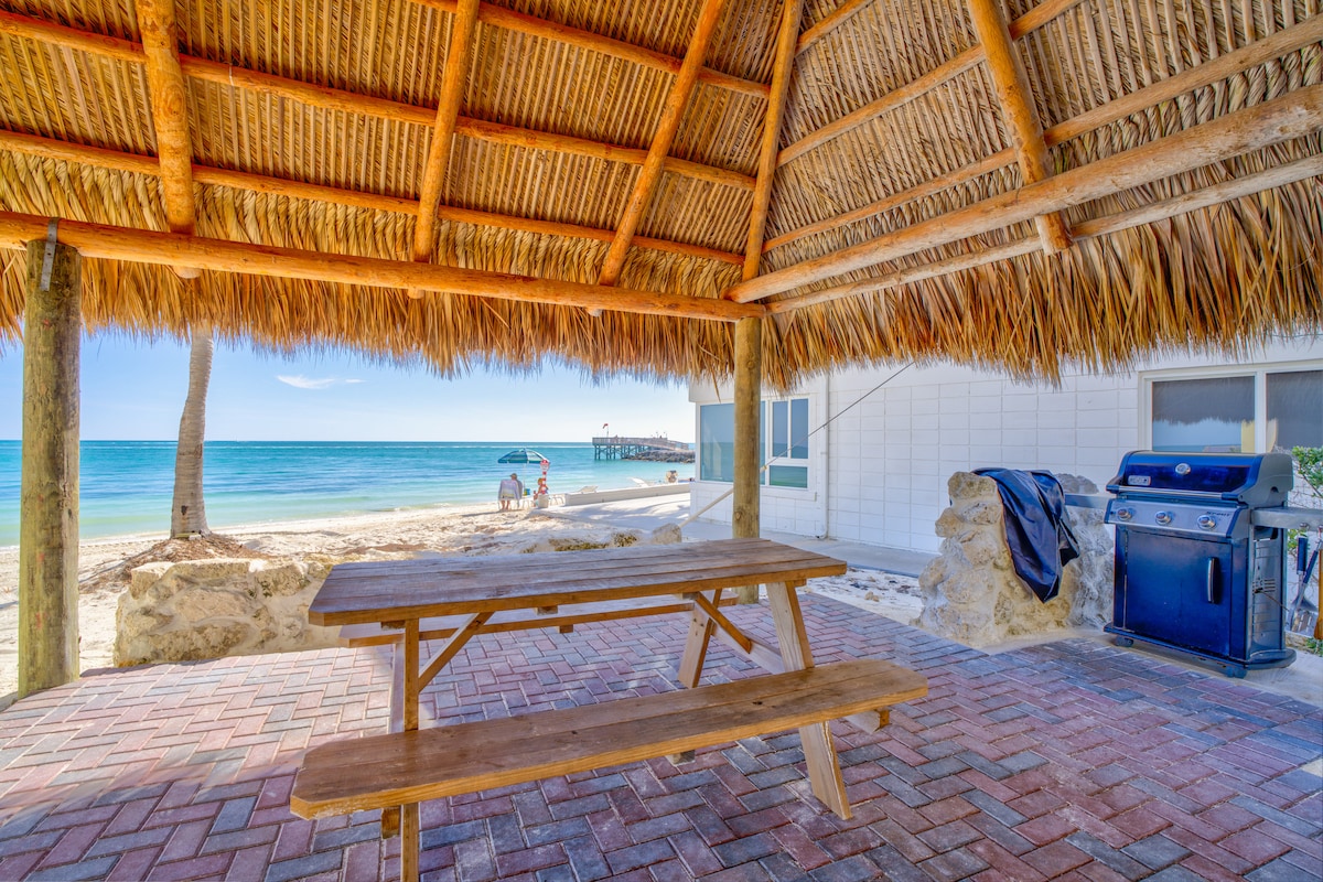 An outdoor dining area with a thatched roof provides shade, featuring a wooden picnic table. In the background, the turquoise waters meet the sandy beach, with a visible pier and beach chairs, while a gas grill is positioned near the stone wall.