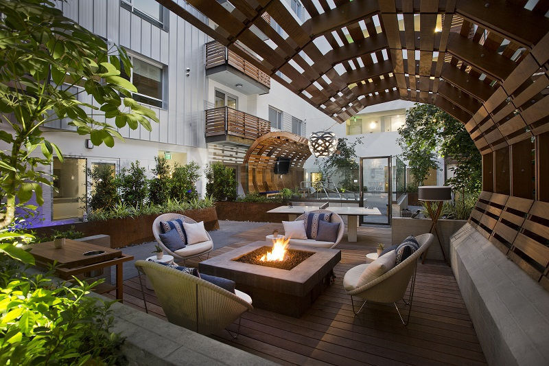 An outdoor communal space is seen featuring a fire pit surrounded by modern seating arrangements. Lush greenery is present, providing natural privacy. The area is enclosed with wood slatted architecture, offering a warm and inviting setting for relaxation and social gatherings.