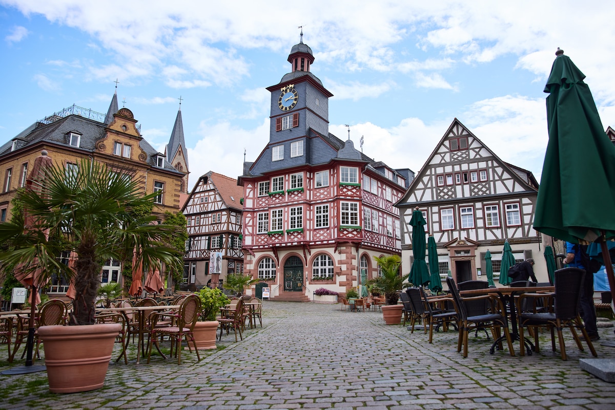 A historic square is framed by traditional half-timbered houses and a tall clock tower. Outdoor seating is arranged around stone-paved pathways, surrounded by lush greenery and palm-like plants. The sky is partly cloudy, adding a serene ambiance to the charming setting.