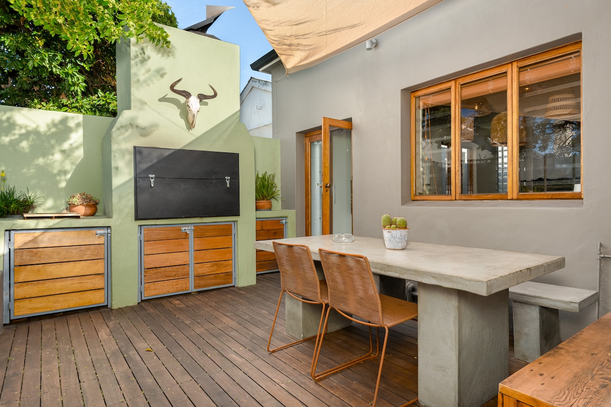 An outdoor patio features a sturdy, rectangular concrete dining table surrounded by four wooden chairs. The area is adorned with greenery and includes a built-in braai for grilling, with a large wall-mounted blackboard behind. Natural light flows through the windows, enhancing the inviting space.