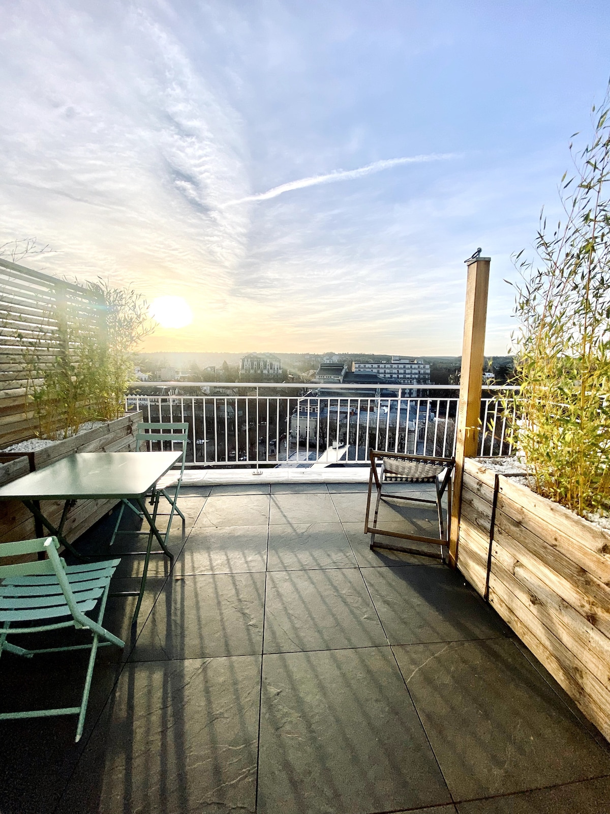 The terrace features a green table and two chairs, surrounded by wooden planters, creating a serene outdoor setting. The sun rises in the background, illuminating the expansive view of the cityscape. A feeling of openness is created by the railing, enhancing the panoramic perspective.