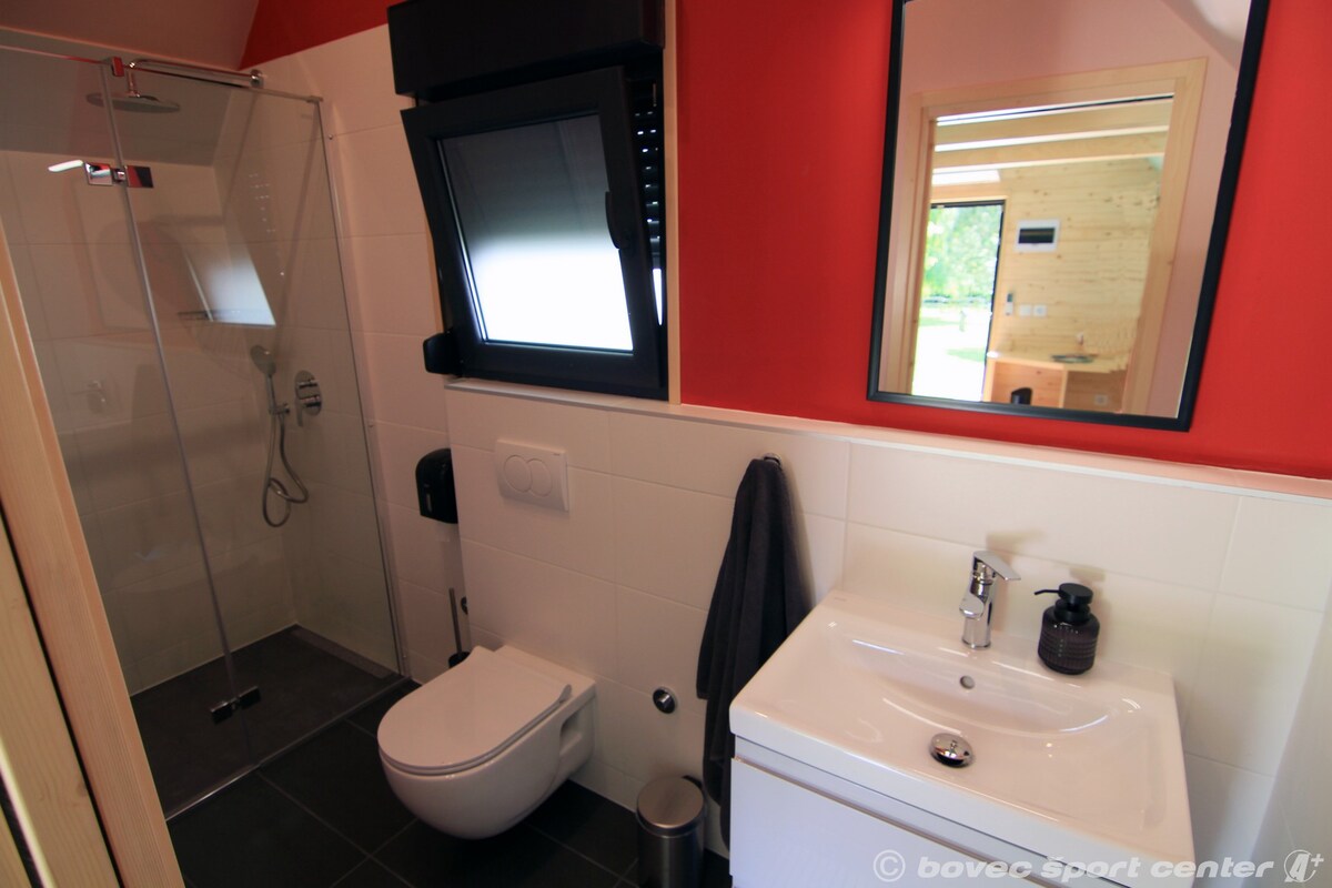 The bathroom features a walk-in shower with transparent glass doors and modern fixtures. A white toilet and sink are positioned against a backdrop of red and neutral tiles. Natural light is provided by a window, complementing the minimalist design.