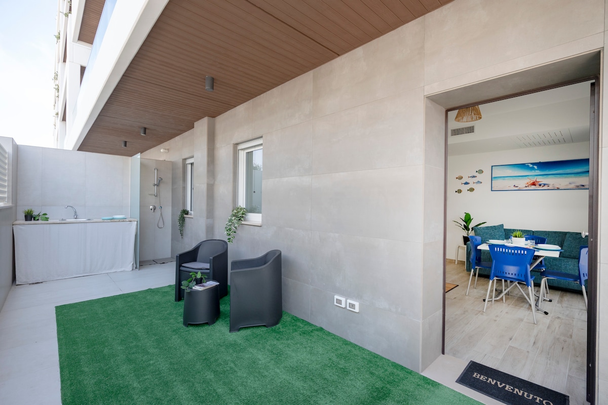 An outdoor area is displayed, featuring a small green lawn. Two black chairs are positioned on the lawn. A doorway leads to a brightly lit interior space with colorful furniture, while a faucet and shower are visible in the corner.