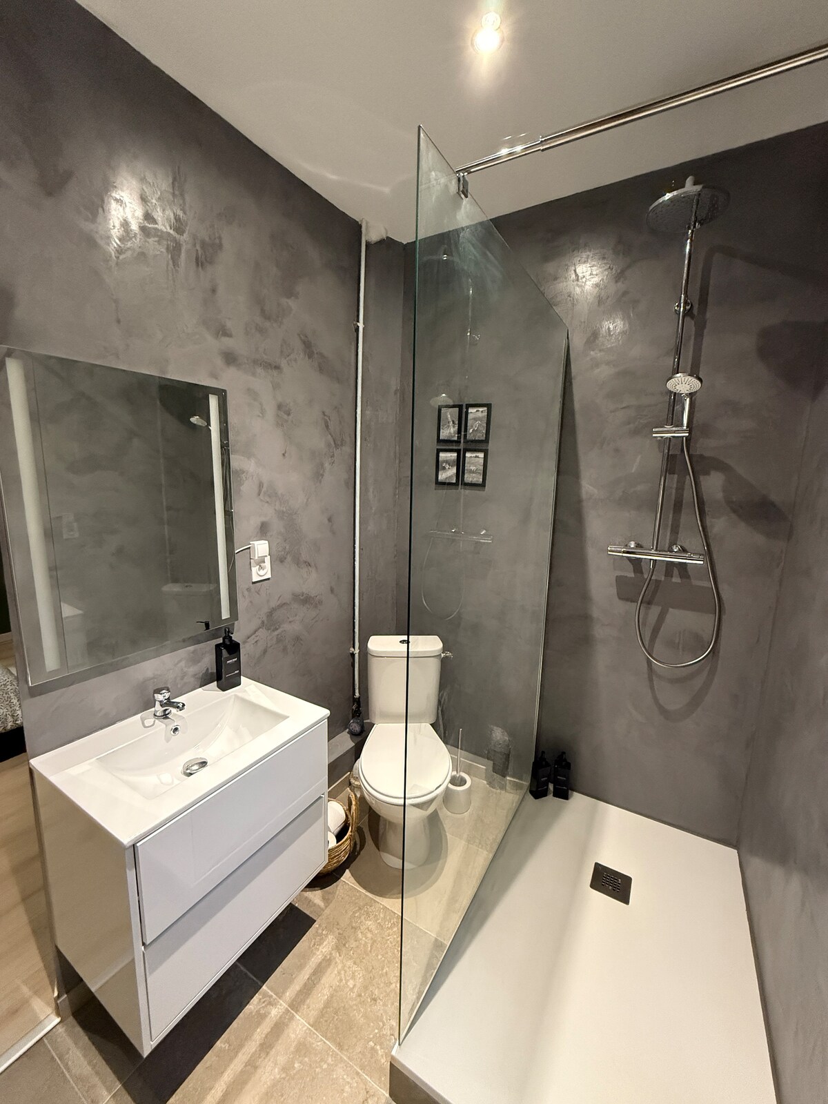 A modern bathroom is presented, featuring a glass shower enclosure and a sleek vanity with a sink. Grey textured walls complement the neutral tones of the floor. A mirror is mounted above the vanity, and minimalist decor is visible on the wall.