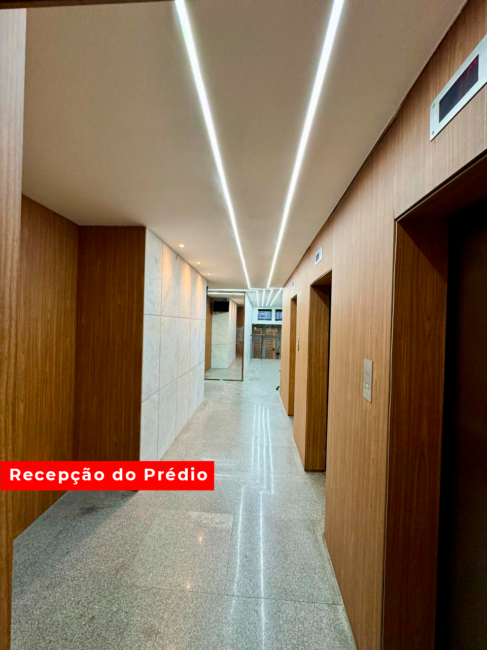 A well-lit corridor features modern wooden paneling and polished stone flooring. The space is enhanced by linear fluorescent lighting along the ceiling. Elevator doors are visible on either side, leading to the building's reception area at the far end.