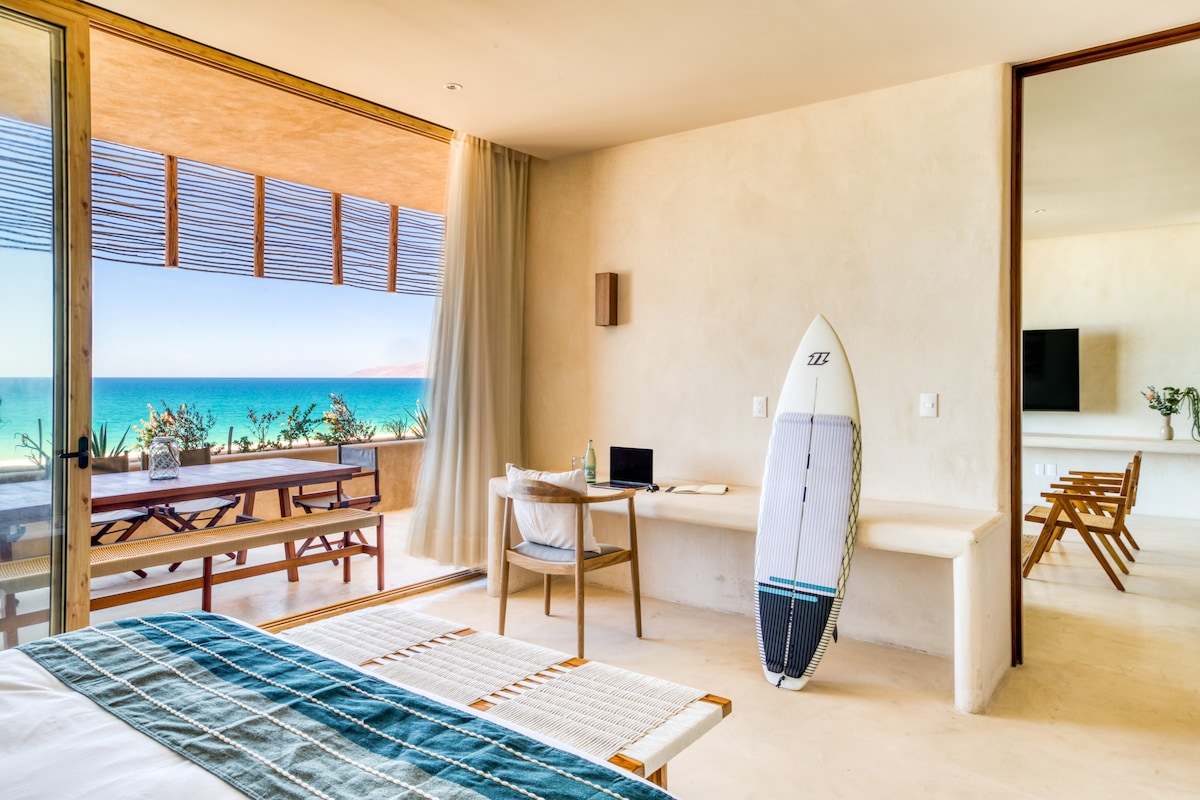 A bright and airy room features a spacious bed with a striped blanket, complemented by a large glass door that provides panoramic views of the ocean. A surfboard leans against a wall near a small desk and chair, while natural light fills the space.