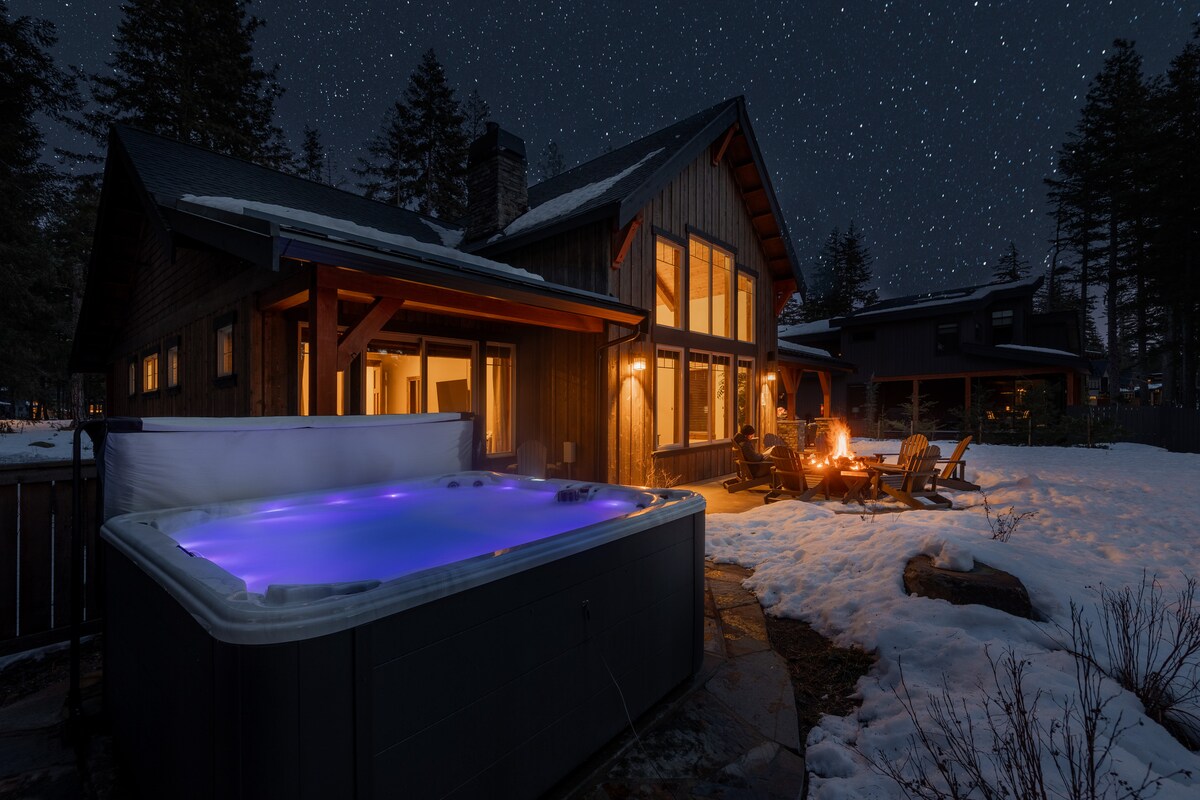 The image captures a serene evening scene featuring a private hot tub with purple lighting and a cozy gas fire pit surrounded by Adirondack chairs. The backdrop showcases a contemporary lodge with large windows, framed by a blanket of snow and a starlit sky.