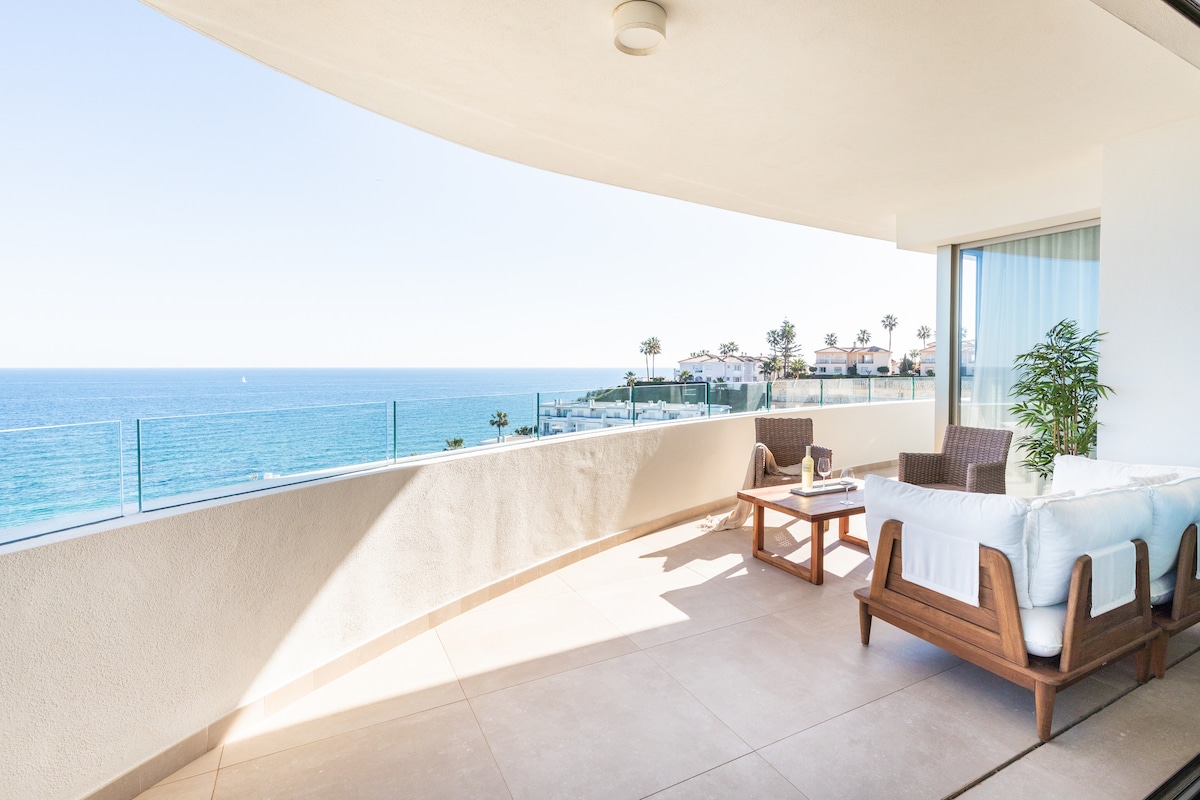 A private terrace offers stunning panoramic views of the ocean, complemented by modern outdoor furniture. Two comfortable chairs and a coffee table are positioned to enjoy the scenery, with a light color palette reflecting the bright coastal environment.