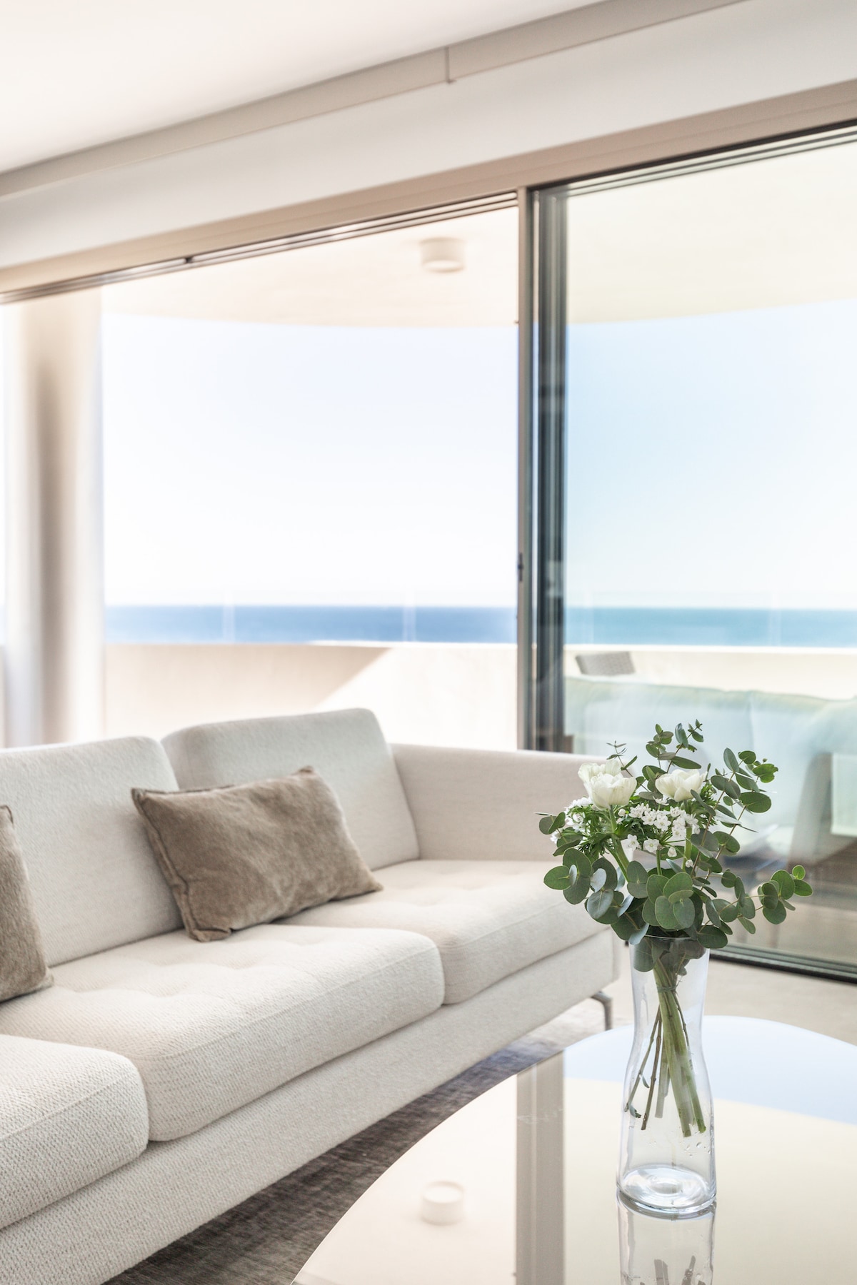 A modern living area features a light-colored sofa with accent cushions, complemented by a glass coffee table. Floor-to-ceiling windows provide unobstructed views of the ocean, allowing natural light to illuminate the space. A vase of fresh greenery adds a touch of elegance.