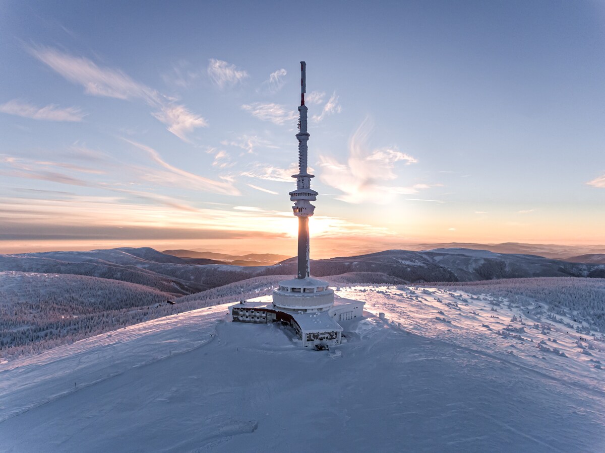 A tall communication tower rises above a snowy landscape, with a vibrant sunset painting the sky in soft hues. The surrounding hills are blanketed in snow, creating a serene winter scene. The vast expanse of snow reflects the warm light of the sunset.