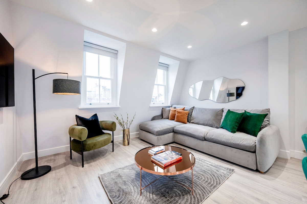 A modern lounge area features a plush grey sofa, an elegant coffee table, and a stylish floor lamp. Large windows invite natural light, while a decorative mirror and potted plants enhance the space. The room is completed by emerald green accent cushions on the sofa.