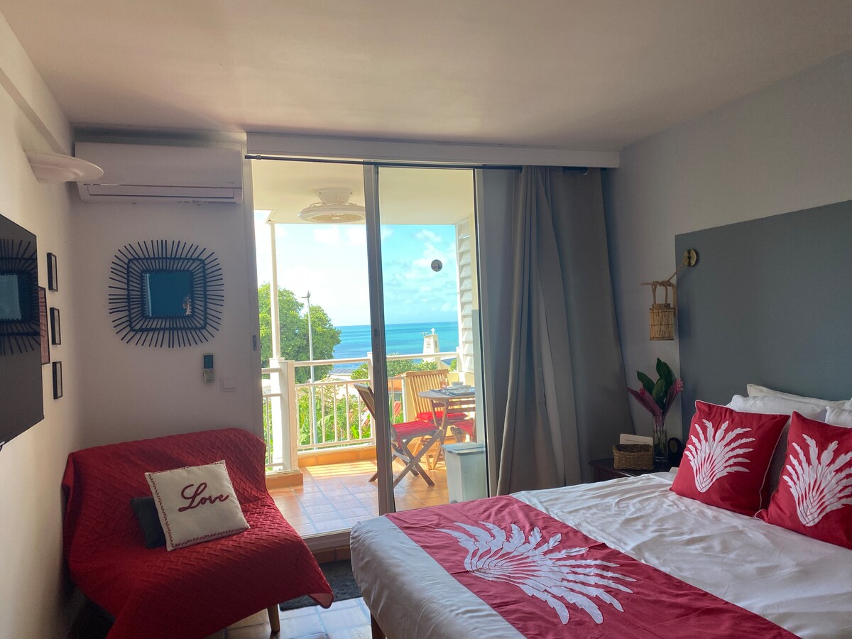 A bright bedroom is shown, featuring a spacious bed with a red and white coverlet. A cozy seating area with a red sofa is visible, complemented by a decorative cushion. Glass doors lead to a balcony with ocean views, allowing natural light to fill the room.