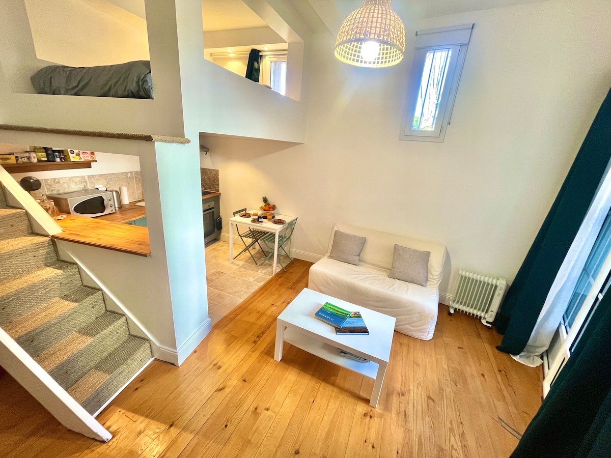 The studio presents an open layout, with a cozy living area featuring a white sofa and a small table set for dining. The kitchenette is visible, alongside a staircase leading to the mezzanine, where a bed is situated. Natural light filters through the windows, enhancing the inviting atmosphere.