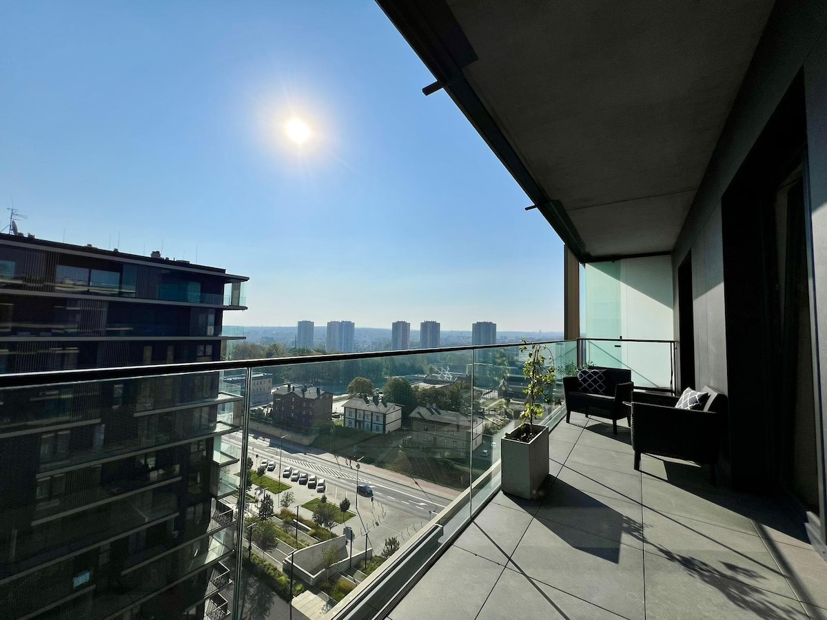 A spacious terrace on the tenth floor features comfortable seating and a potted plant. A clear view of the skyline extends in the distance, with buildings positioned along a clean city road under a bright sun.