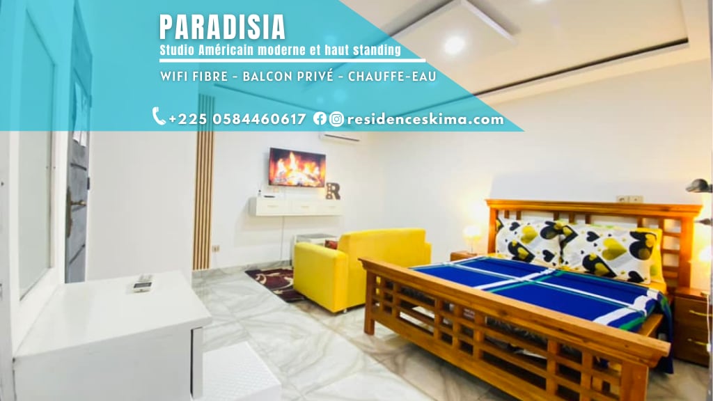 A modern studio features a wooden bed with a blue and green blanket. A yellow sofa is positioned nearby, alongside a wall-mounted television. The room is designed with neutral tones, and light-colored tiles enhance the spaciousness of the area.