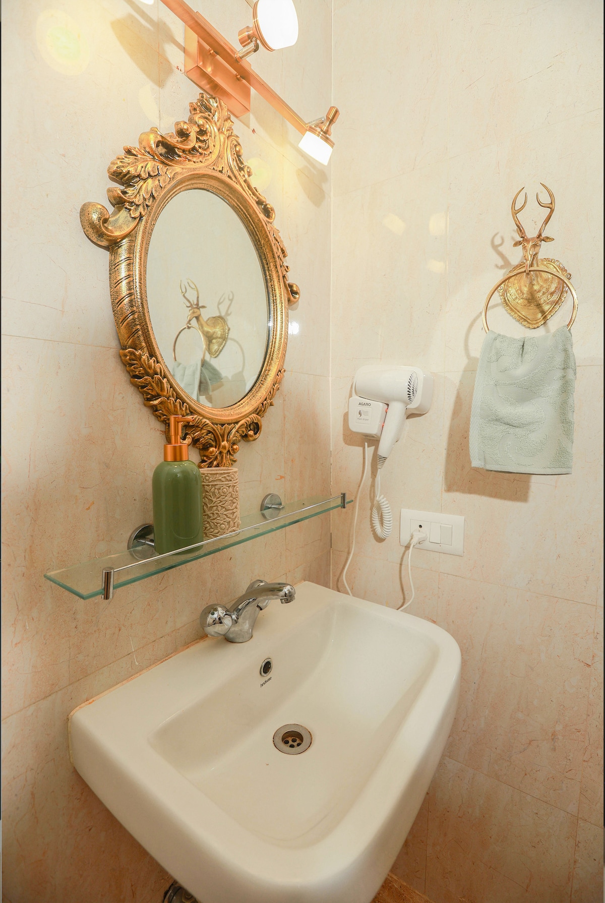 A compact bathroom space features a wall-mounted sink with a modern faucet. A decorative mirror with a golden frame is positioned above the sink. A hand towel hangs from a stylish holder, accompanied by a soap dispenser and a hairdryer on the wall beside it.