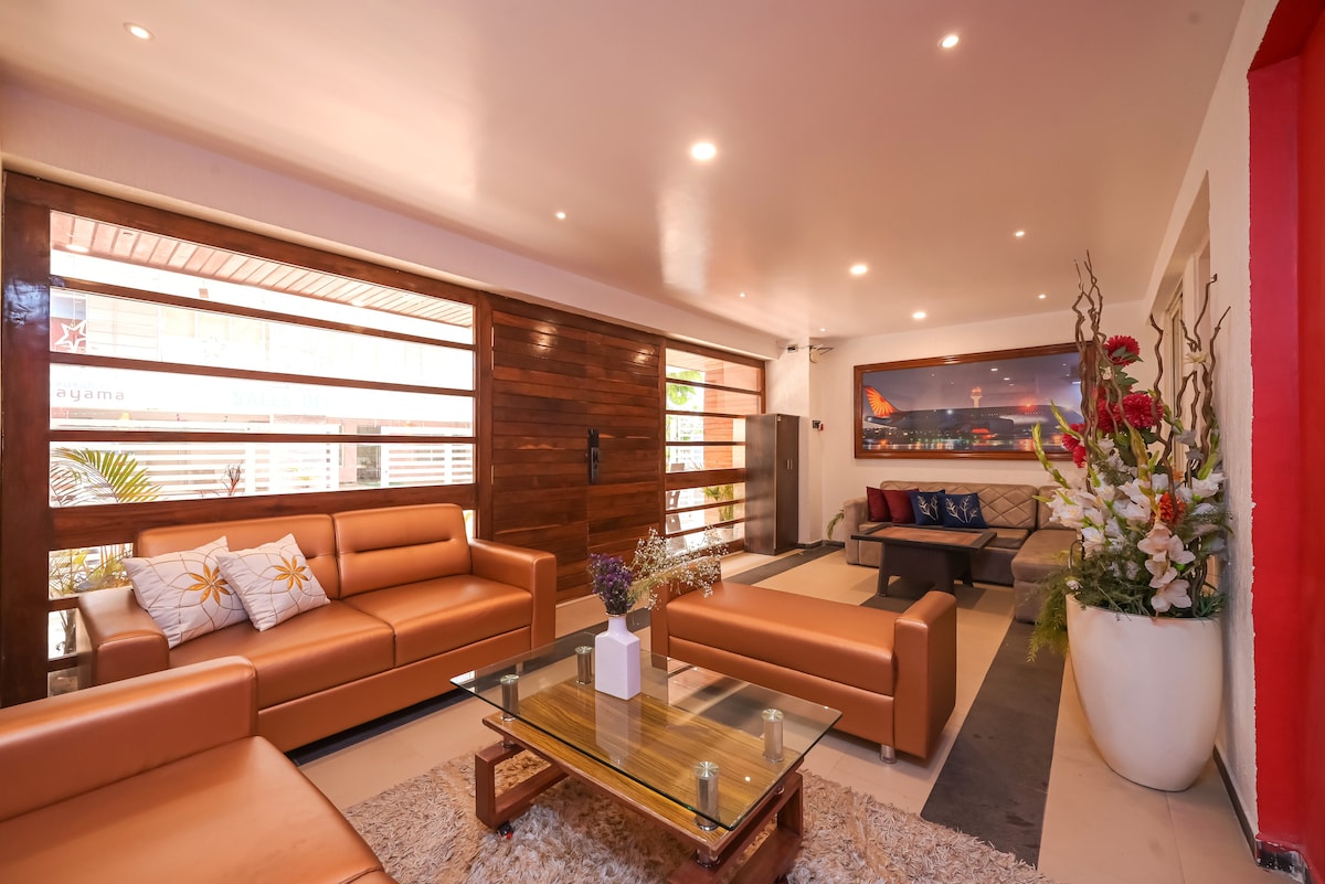 A well-furnished living room features large glass windows allowing natural light to fill the space. Comfortable sofas are arranged around a central coffee table, while decorative plants and flowers add a touch of elegance. Art pieces adorn the walls, enhancing the room's modern design.