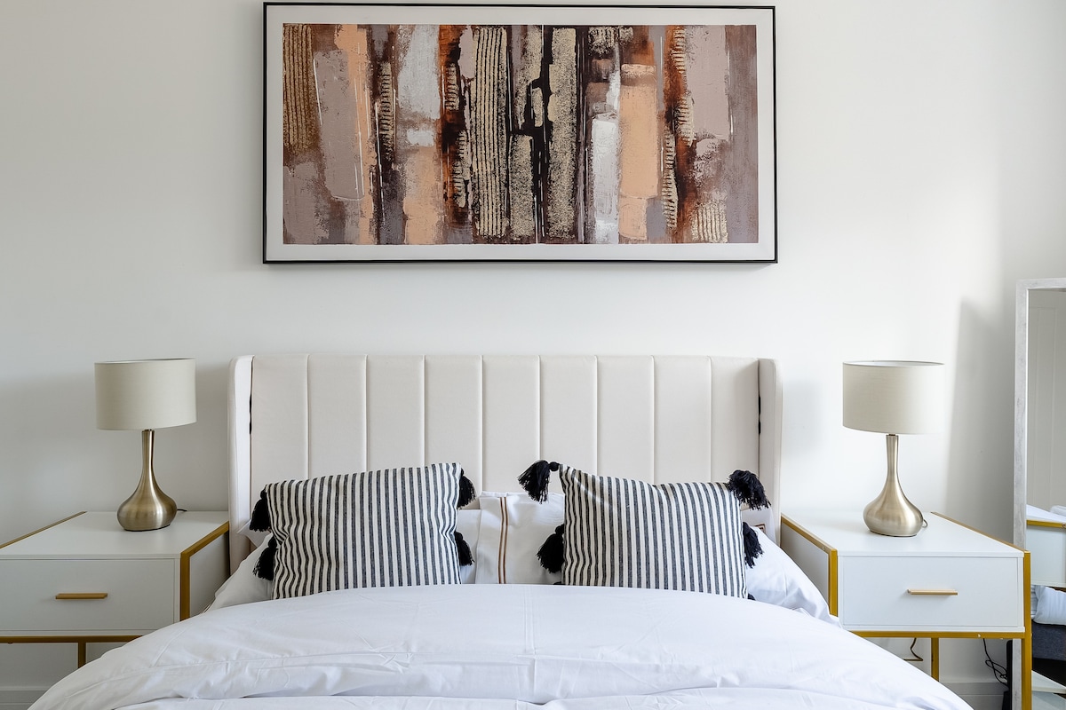 A modern bedroom features a spacious bed with smooth white linens and striped accent pillows. Flanking the bed are two nightstands topped with stylish lamps. Above the bed, an abstract artwork adds color to the neutral walls, while a full-length mirror is positioned nearby.