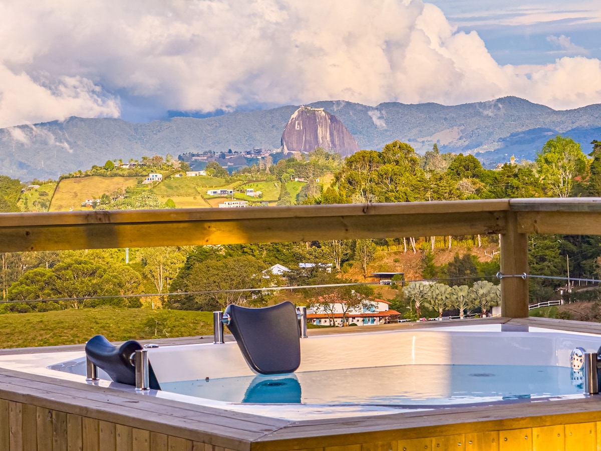 A private jacuzzi sits on a wooden deck, overlooking rolling hills and a prominent rock formation in the distance. The landscape features lush greenery under a partly cloudy sky, creating a serene environment for relaxation and enjoying nature.