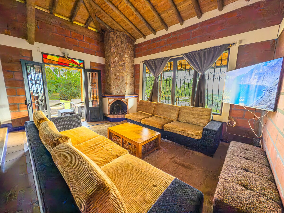 A spacious living area features a cozy seating arrangement with a large, textured sofa. Natural light streams through multiple windows adorned with curtains, highlighting the stone fireplace and decorative stained glass. A flat-screen television is mounted on the wall, providing a modern touch to the rustic decor.