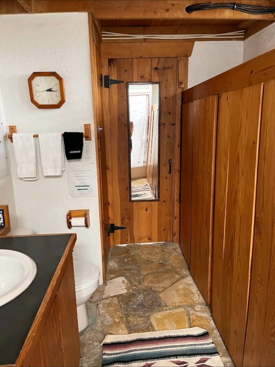 A wooden bathroom is presented with stone flooring, featuring a circular mirror above a dark counter. Soft towels are neatly hung beside the door, which has a small mirror on its surface. Natural light filters in through a window at the end of the space.