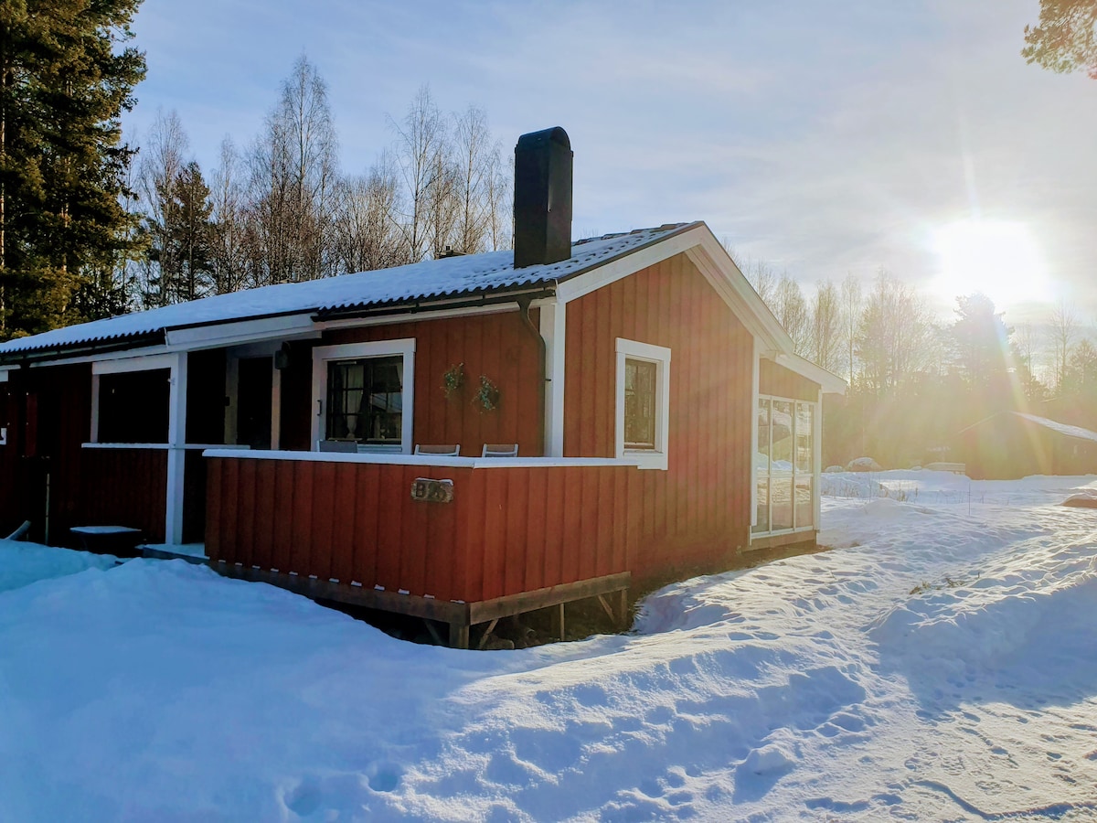 A charming red cottage is depicted, partially surrounded by snow. The structure features white-framed windows and a sloped roof. Sunlight is visible in the background, creating a serene winter atmosphere. A chimney is situated on top, hinting at warmth inside.