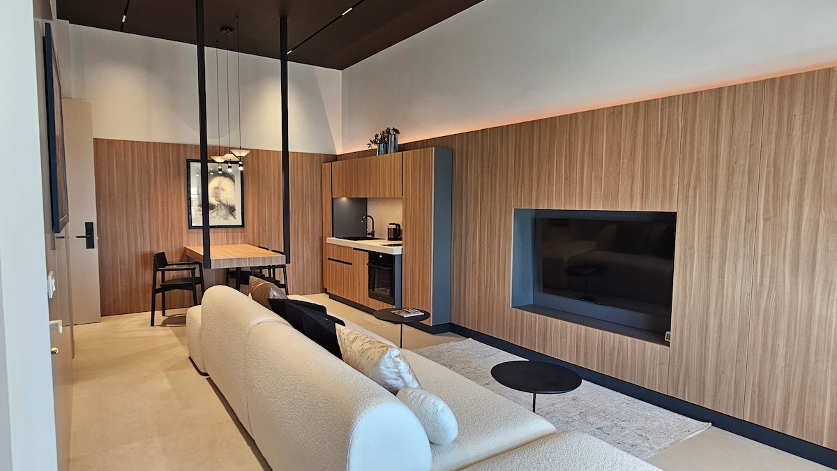 A modern living space is presented with light wood paneling and a minimalist design. A spacious sofa faces a wall-mounted television, while a dining table and kitchenette are visible in the background, enhancing the functionality of the area. Large windows allow natural light to fill the room.