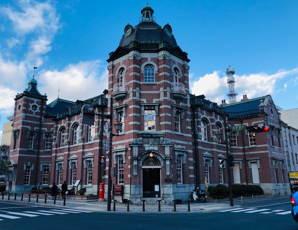 A historic red brick building features a unique architectural design with a domed tower and intricate detailing. Large windows and stone accents complement the facade. The structure is surrounded by neatly arranged landscaping and is positioned prominently against a clear blue sky.