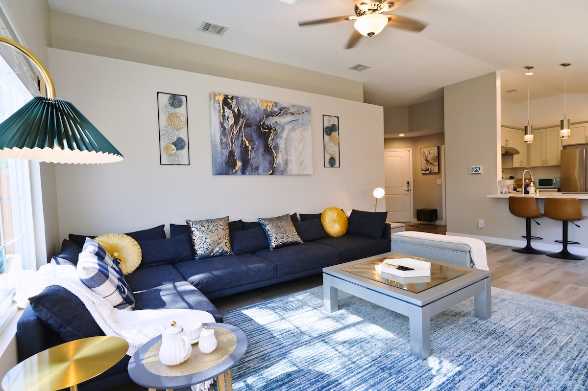 A spacious living area is furnished with a large sectional sofa adorned with decorative cushions. A coffee table sits in the center, and a ceiling fan provides air circulation. Art pieces decorate the walls, while a well-equipped kitchen is visible in the background.