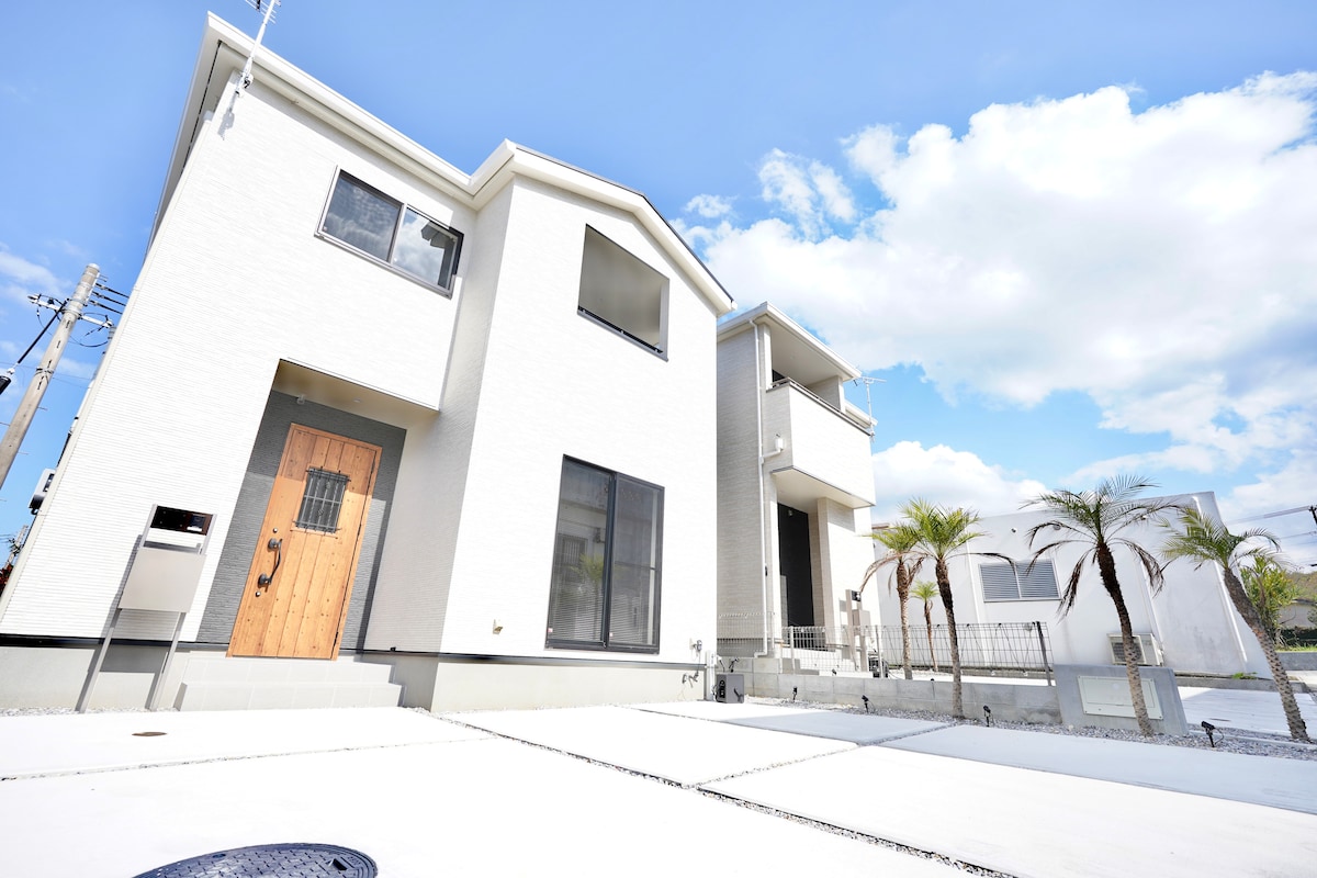The exterior of the newly built hotel features a modern design with a white facade and large windows. A wooden front door leads to the entrance, while palm trees add a touch of greenery to the landscape. Bright blue skies complete the inviting atmosphere.