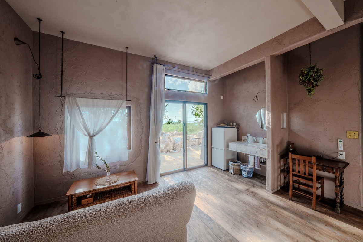 A light and airy living area features a seating area with a soft couch and a wooden coffee table. Natural light enters through a large glass door that opens to the outdoor space, complemented by a simple vanity and sink area against textured walls.