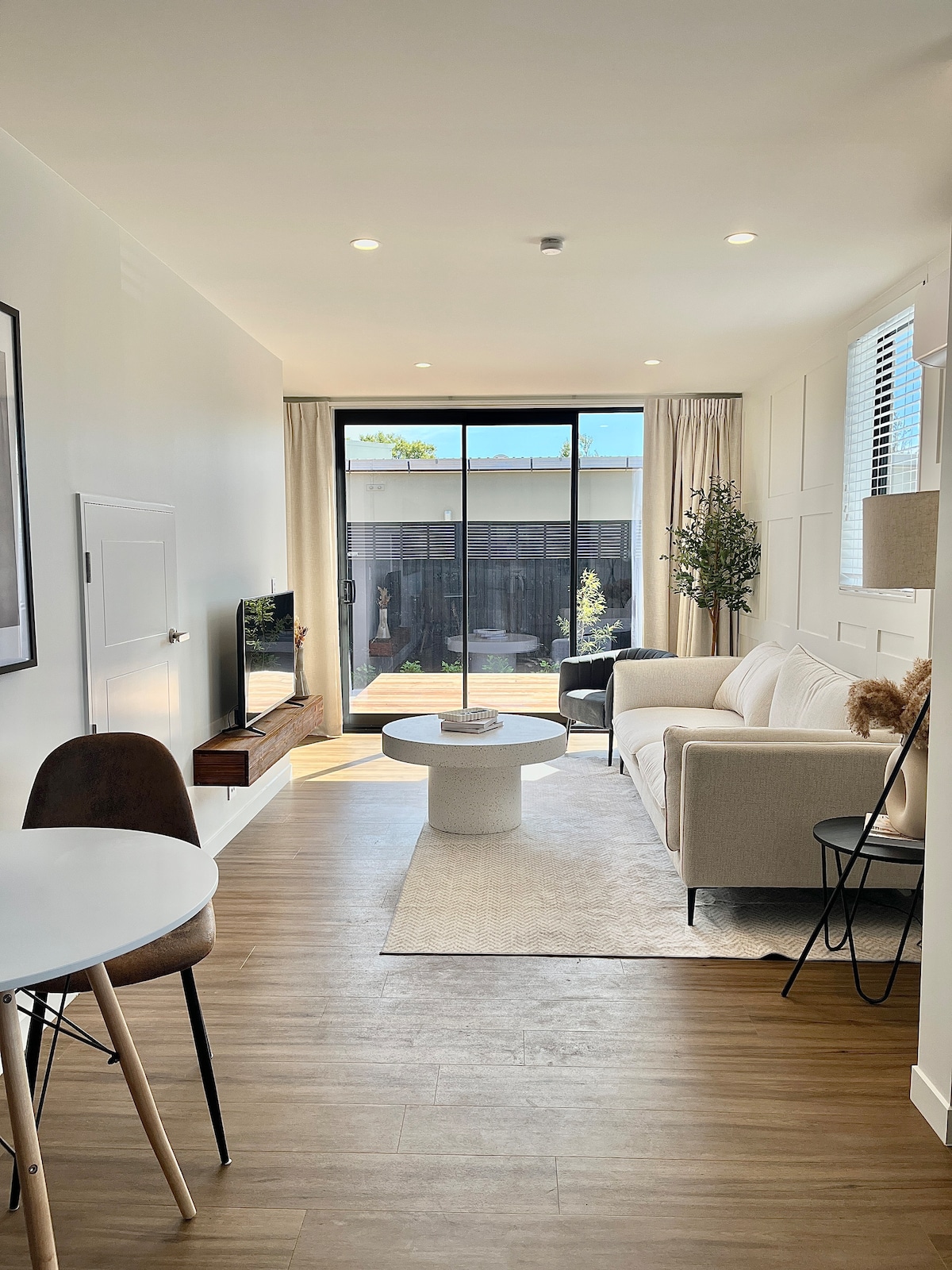 A light-filled lounge area features a comfortable sofa and a modern coffee table. A television is mounted on the wall across from the seating. Large glass doors provide access to the outdoor courtyard, allowing natural light to enhance the space. Neutral decor creates a calming atmosphere.