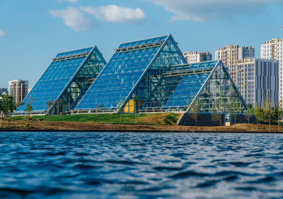 A modern glass structure features a series of triangular roofs, reflecting natural light and surrounded by green landscaping. In the foreground, a serene body of water mirrors the architecture, with urban buildings visible in the background under a clear blue sky.