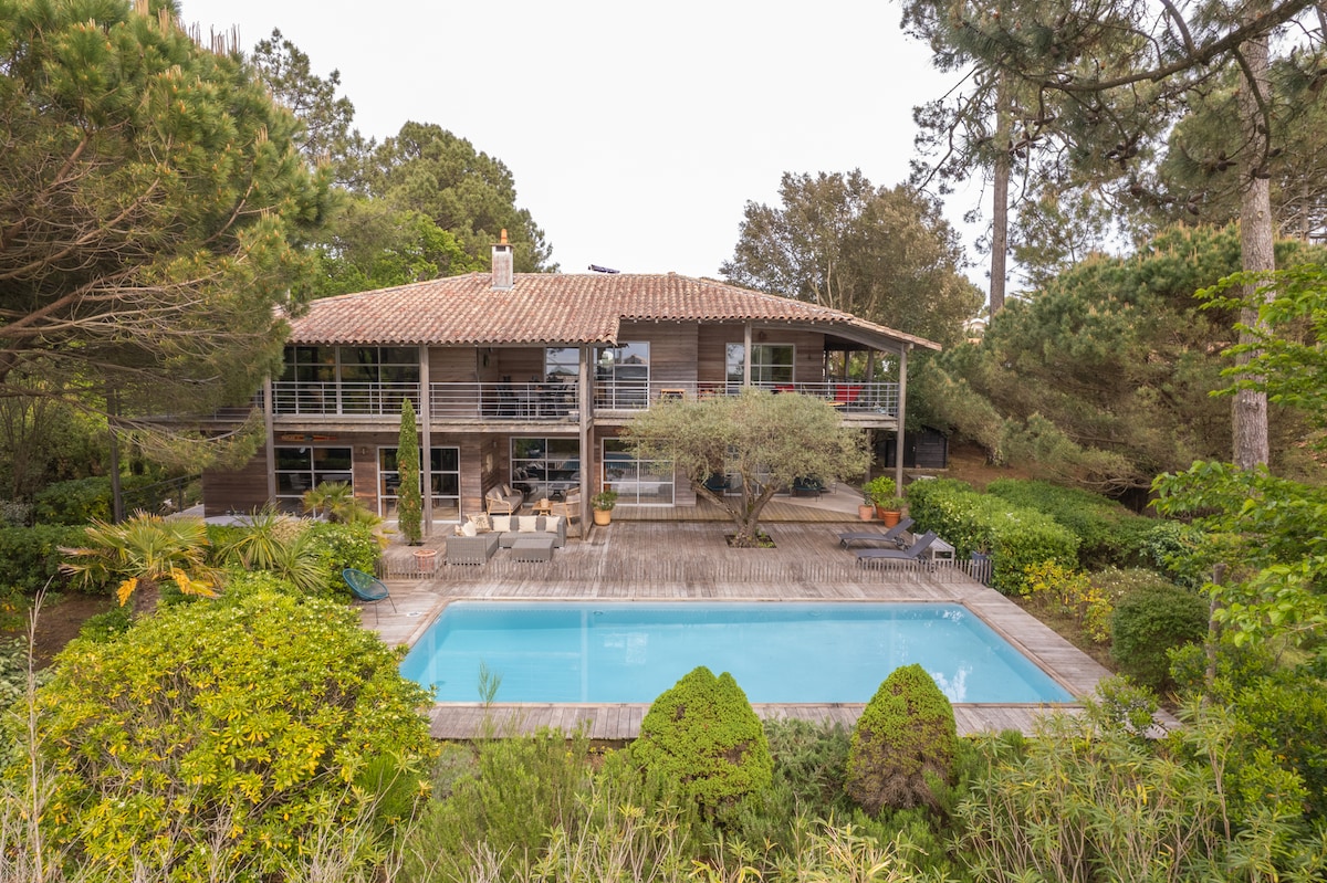 A modern villa constructed with wood features large glass doors, blending with the surrounding greenery. An inviting pool is situated in front of the house, surrounded by a landscaped garden, with outdoor seating areas visible on the terrace.