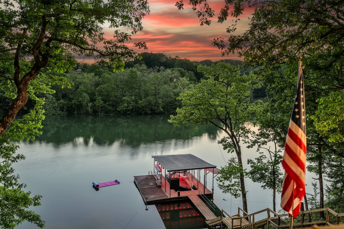 A private dock extends into a calm lake, surrounded by lush green trees reflecting in the water. A colorful sunset casts a warm glow across the sky. An American flag gently flutters on a nearby post, enhancing the serene lakeside scene.