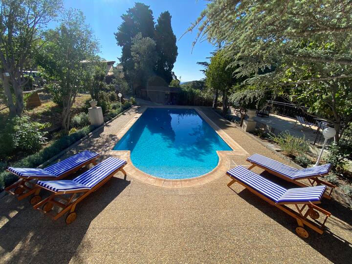 A large swimming pool is surrounded by a sun deck featuring several wooden loungers with blue and white striped cushions. Lush greenery and trees are visible in the background, enhancing the tranquil outdoor space.