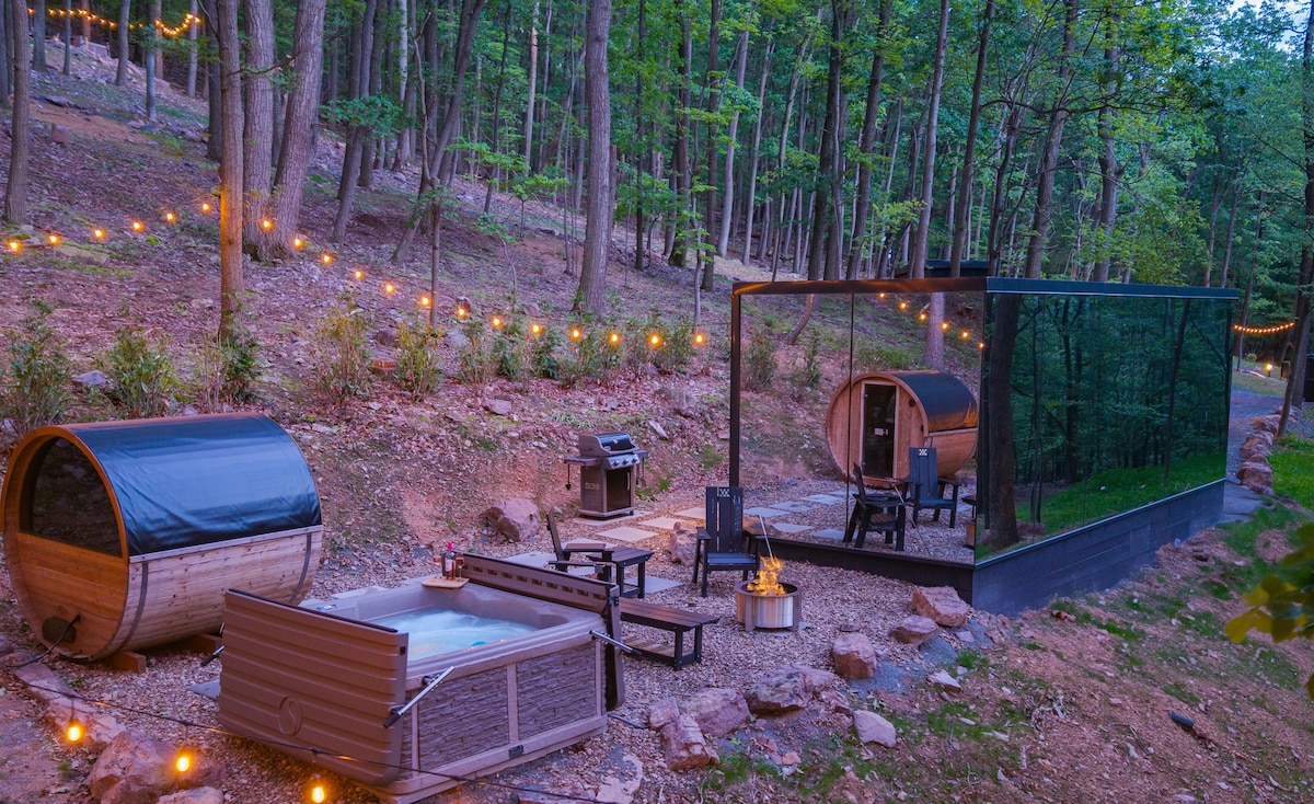 The outdoor area features a hot tub and a private sauna presented in modern barrel designs. A fire pit surrounded by seating creates a cozy gathering space. String lights are visible among the trees, enhancing the serene atmosphere of the wooded surroundings.